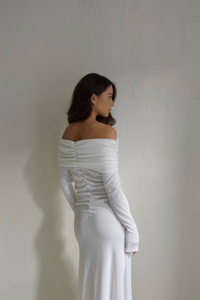 A woman, with her back and left side facing the camera, models an off-the-shoulder, long-sleeved Dalia Dress that showcases gathered detailing along the back. She has long, wavy brown hair and stands against a plain, light-colored wall.