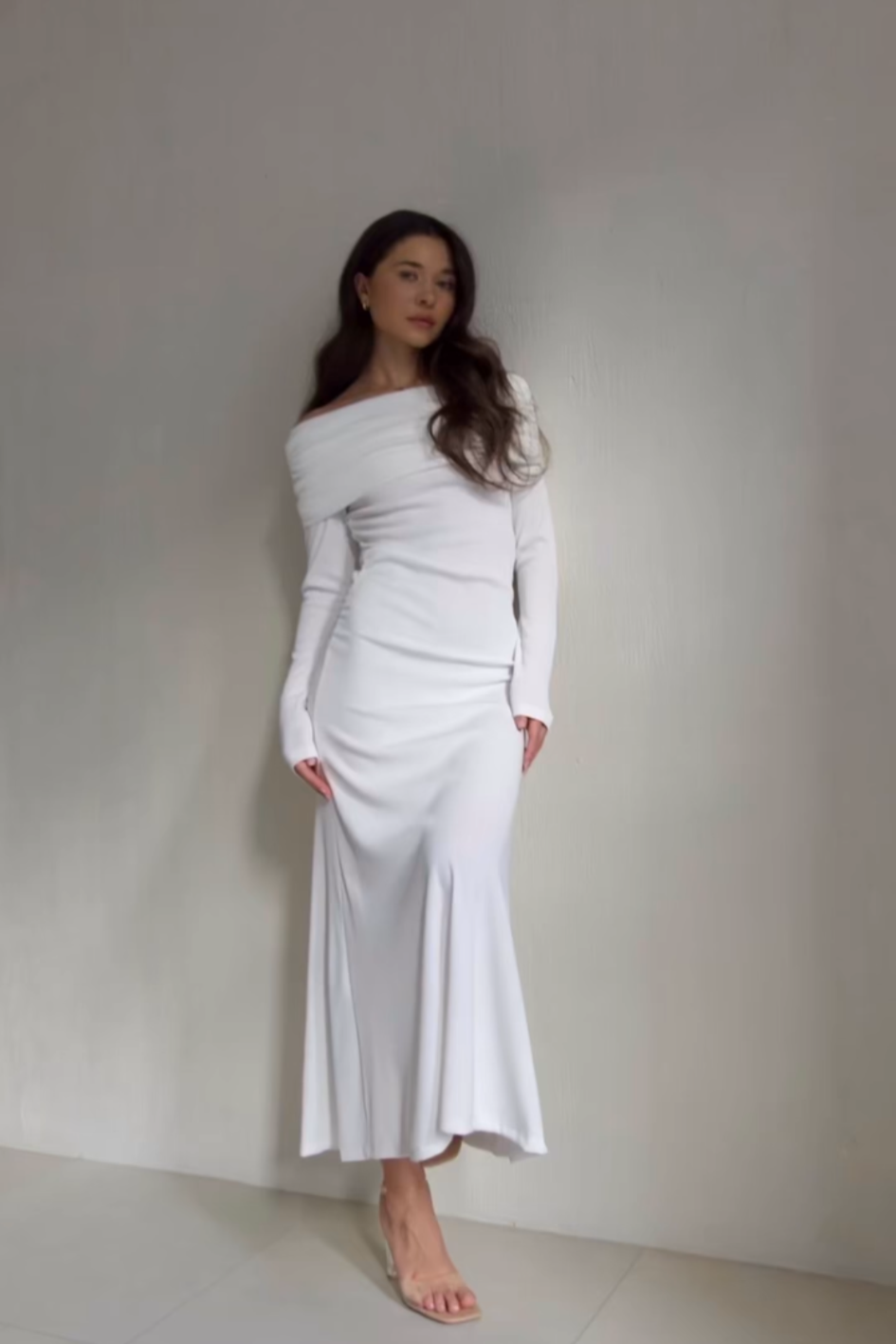 A woman stands against a plain light-colored wall, adorned in the Dalia Dress—an elegant off-shoulder design that gracefully extends to her ankles. Her long, wavy hair cascades over her shoulders as she complements the outfit with nude strappy heeled sandals. She gazes at the camera with a subtle, relaxed pose, epitomizing a fashion-forward wardrobe.