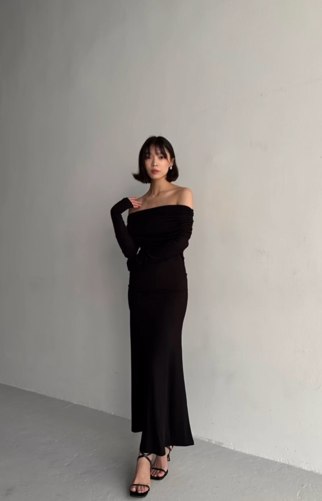 Against a plain light gray wall, a woman with shoulder-length dark hair emanates elegance in a long, off-shoulder Dalia Dress featuring long sleeves. She completes her stylish ensemble with black strappy heels and gazes at the camera while resting one hand on her chest, epitomizing a fashion-forward wardrobe.