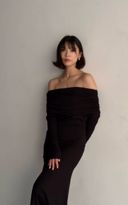 A woman with short, dark hair stands against a light-colored wall. She is wearing a long Dalia Dress with an off-the-shoulder design, her hands resting casually. She looks directly at the camera with a neutral expression, exuding confidence in her fashion-forward wardrobe.