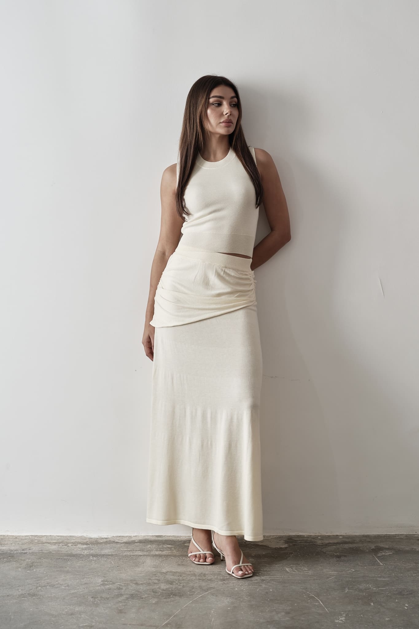 Model wearing a white Andrea Set featuring a sleeveless knit top with gathered detailing and a matching maxi skirt, styled with white strappy sandals, posing against a grey and white textured background