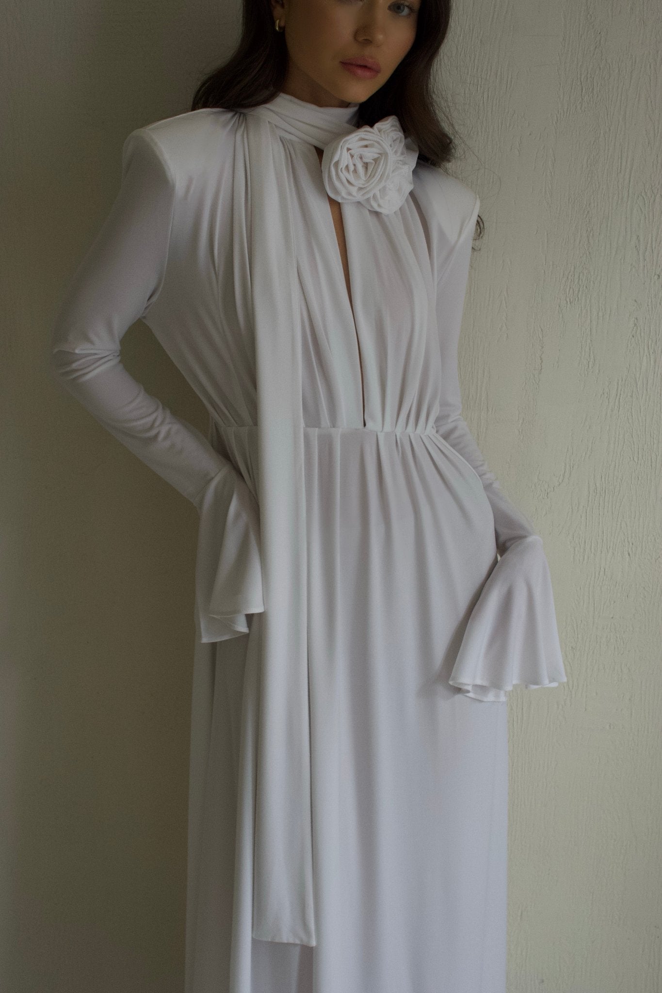 A person wearing an Evey dress, a flowing white gown with long sleeves and flared cuffs, stands against a textured wall. Ideal for special occasions, the dress features a scarf-like detail around the neck with a rosette embellishment. The person's face is partially visible.