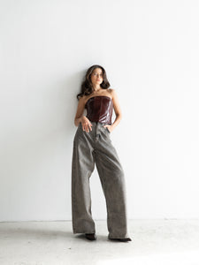 A woman with long dark hair leans against a white wall, exuding effortless grace. She is wearing the timeless essential Imari Corset in burgundy paired with high-waisted, wide-leg gray jeans. Her posture is relaxed, one leg slightly bent and her gaze directed to the side.