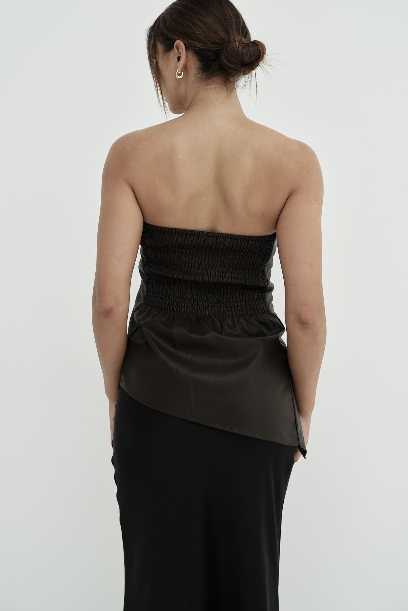 A woman stands against a plain background, modeling the Ellie leather top. The strapless design features a pleated dark green bodice paired with a sleek black skirt. Her dark hair is styled back, and she's completed the look with black heels, adopting a relaxed pose with one hand placed behind her back.