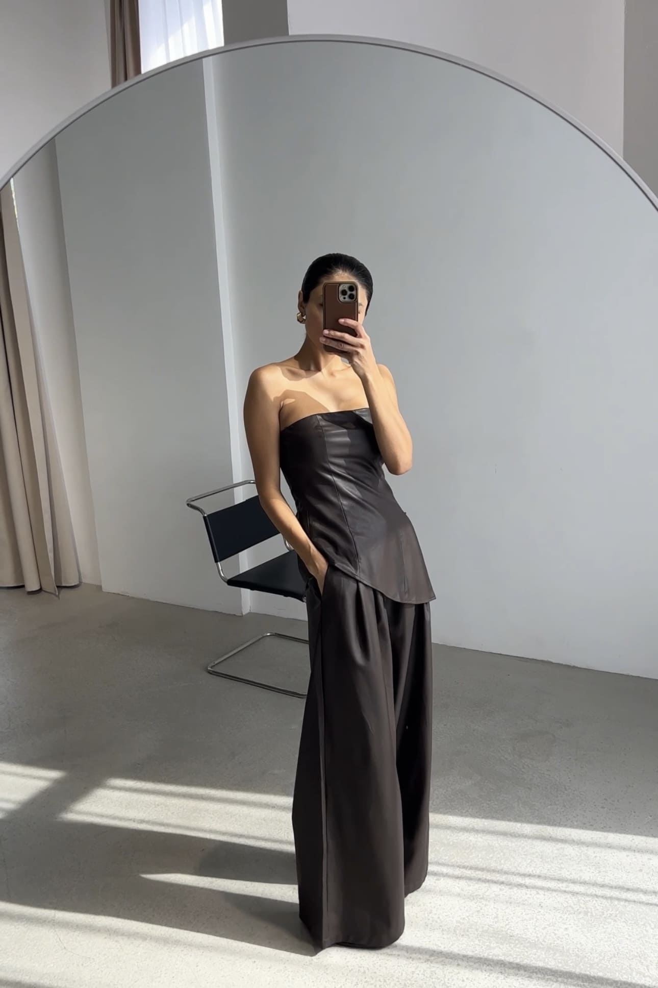 A person stands in front of a large mirror, taking a selfie with a smartphone while wearing the Ellie leather top. The strapless black outfit features wide-leg pants and a pleated texture. A chair and curtains are visible in the background, enhancing the stylish ambiance.