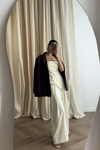 A person stands in front of a mirror, wearing Ellie leather top with a strapless, pleated texture and a black blazer draped over their shoulders. They are holding a smartphone, and beige curtains hang softly in the background.