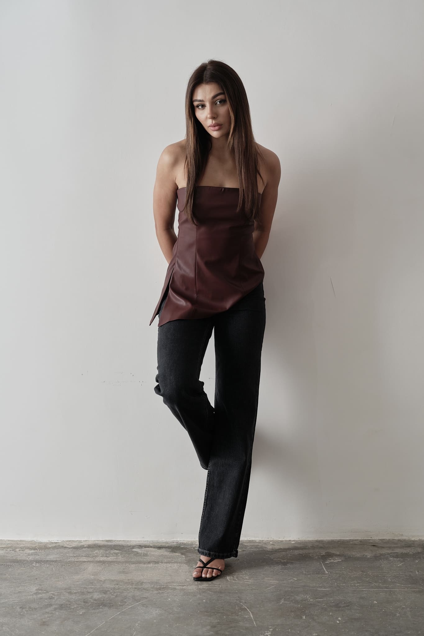 A woman stands against a plain background, modeling the Ellie leather top. The strapless design features a pleated dark green bodice paired with a sleek black skirt. Her dark hair is styled back, and she's completed the look with black heels, adopting a relaxed pose with one hand placed behind her back.