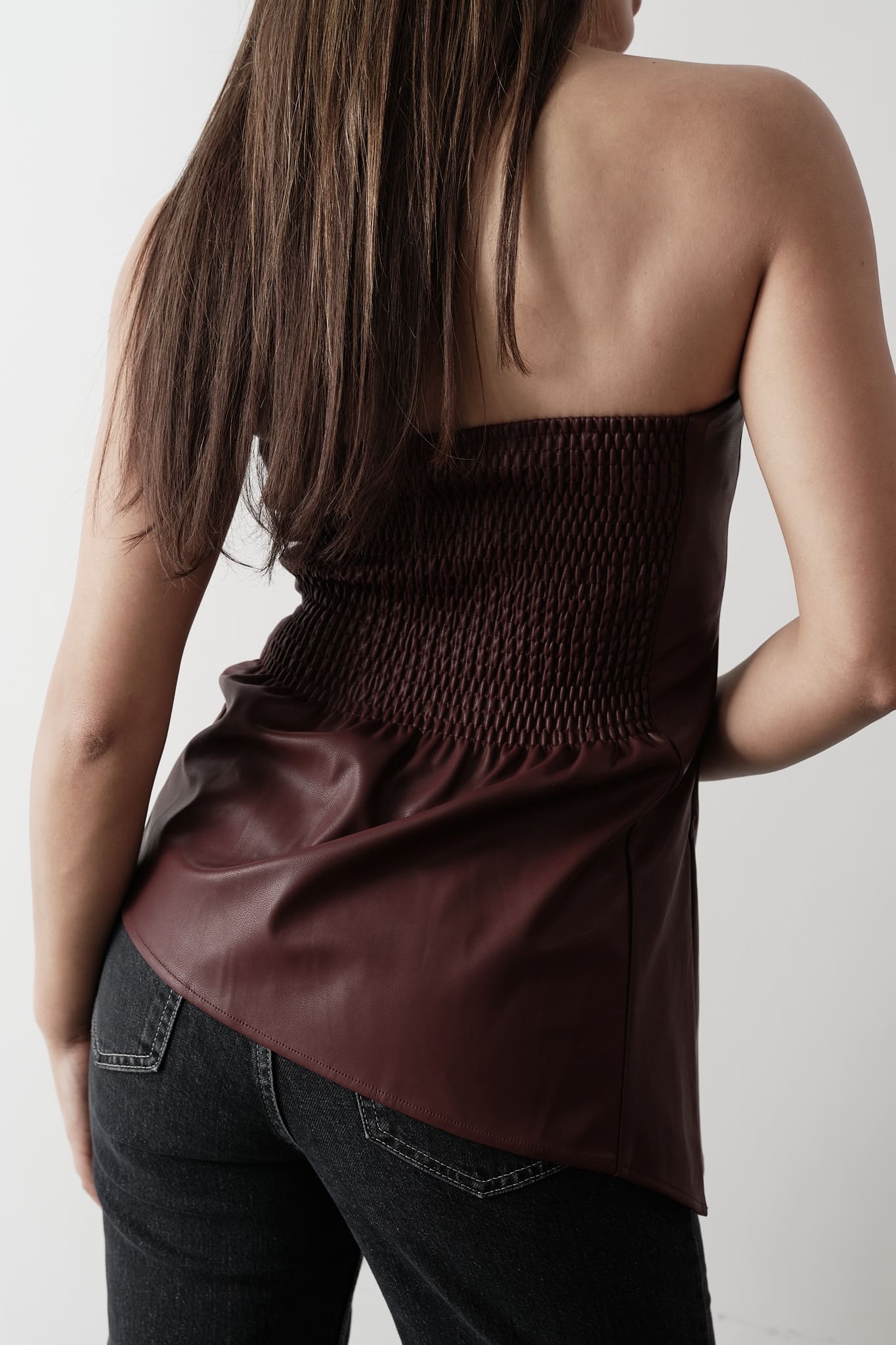 A woman stands against a plain background, modeling the Ellie leather top. The strapless design features a pleated dark green bodice paired with a sleek black skirt. Her dark hair is styled back, and she's completed the look with black heels, adopting a relaxed pose with one hand placed behind her back.