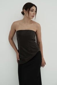 A woman with dark hair styled in an updo wears Ellie leather top, featuring a pleated texture. Positioned against a plain white background, she gazes to the side with one hand resting on her hip, showcasing the elegance of its butter-soft knit fabric.