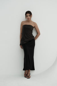 A woman stands against a plain background, modeling the Ellie leather top. The strapless design features a pleated dark green bodice paired with a sleek black skirt. Her dark hair is styled back, and she's completed the look with black heels, adopting a relaxed pose with one hand placed behind her back.