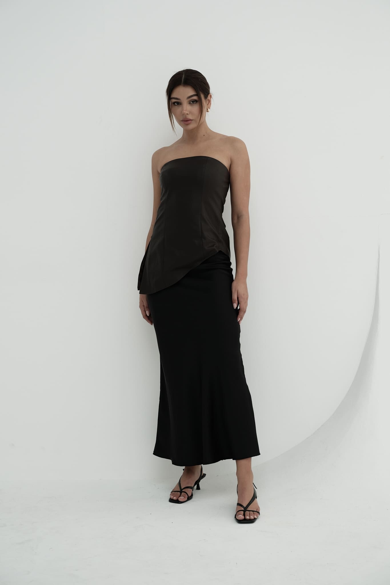 A woman stands against a plain background, modeling the Ellie leather top. The strapless design features a pleated dark green bodice paired with a sleek black skirt. Her dark hair is styled back, and she's completed the look with black heels, adopting a relaxed pose with one hand placed behind her back.