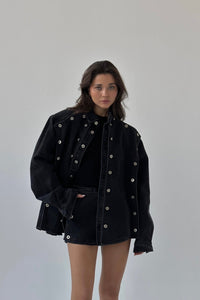 A person with long dark hair is wearing an oversized black Etta set jacket with metallic button details over a black outfit. The jacket features silver grommets, adding an edgy touch. The person is standing against a plain white background, looking slightly to the side with a neutral expression.