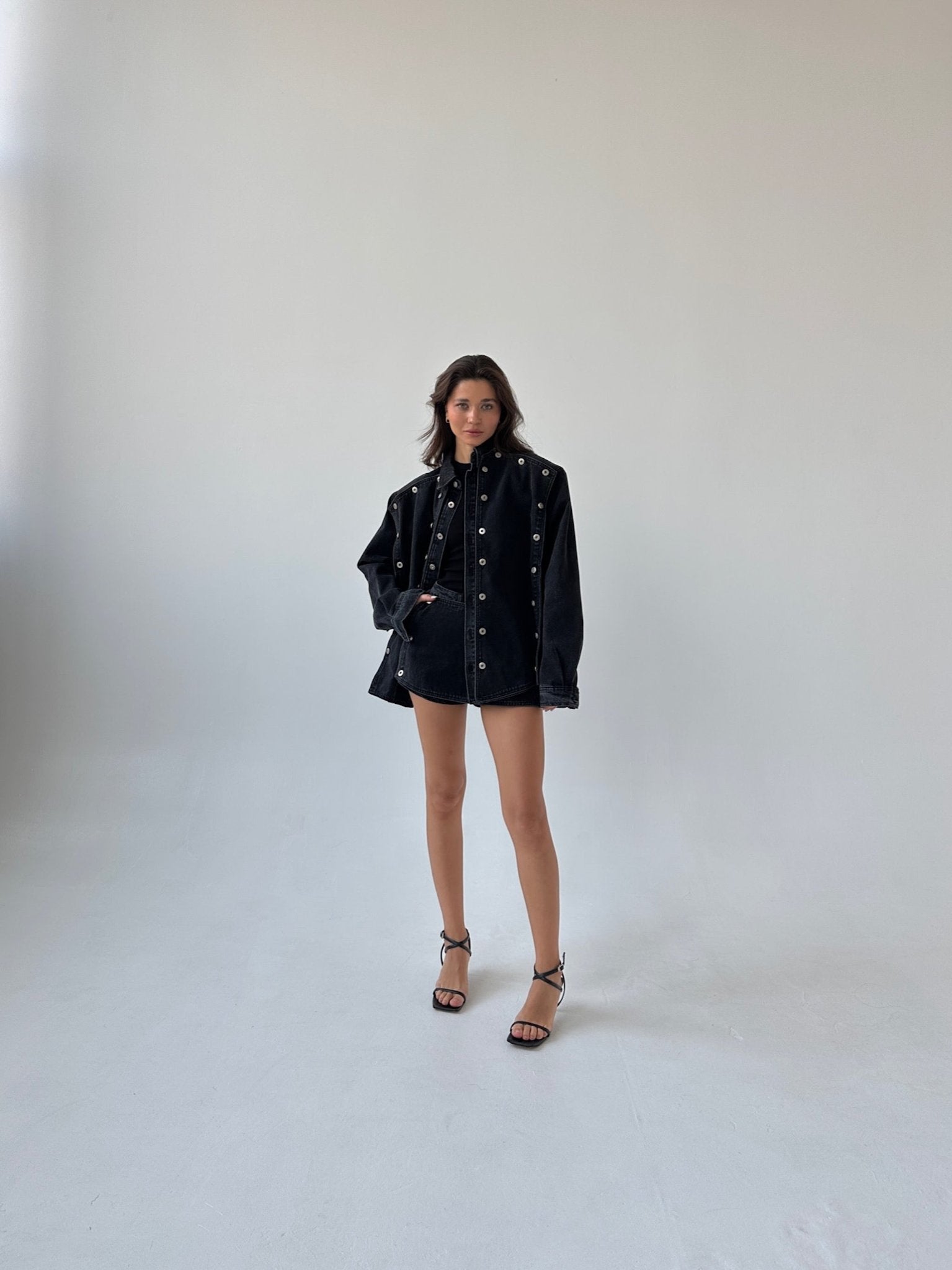 A woman stands in a minimalist studio with a white background, wearing the stylish Etta set, which features an oversized cropped black denim jacket adorned with silver grommets and matching shorts. She is also wearing black strappy high heels and has long, wavy hair.