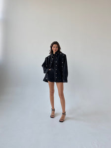 A woman stands in a minimalist studio with a white background, wearing the stylish Etta set, which features an oversized cropped black denim jacket adorned with silver grommets and matching shorts. She is also wearing black strappy high heels and has long, wavy hair.