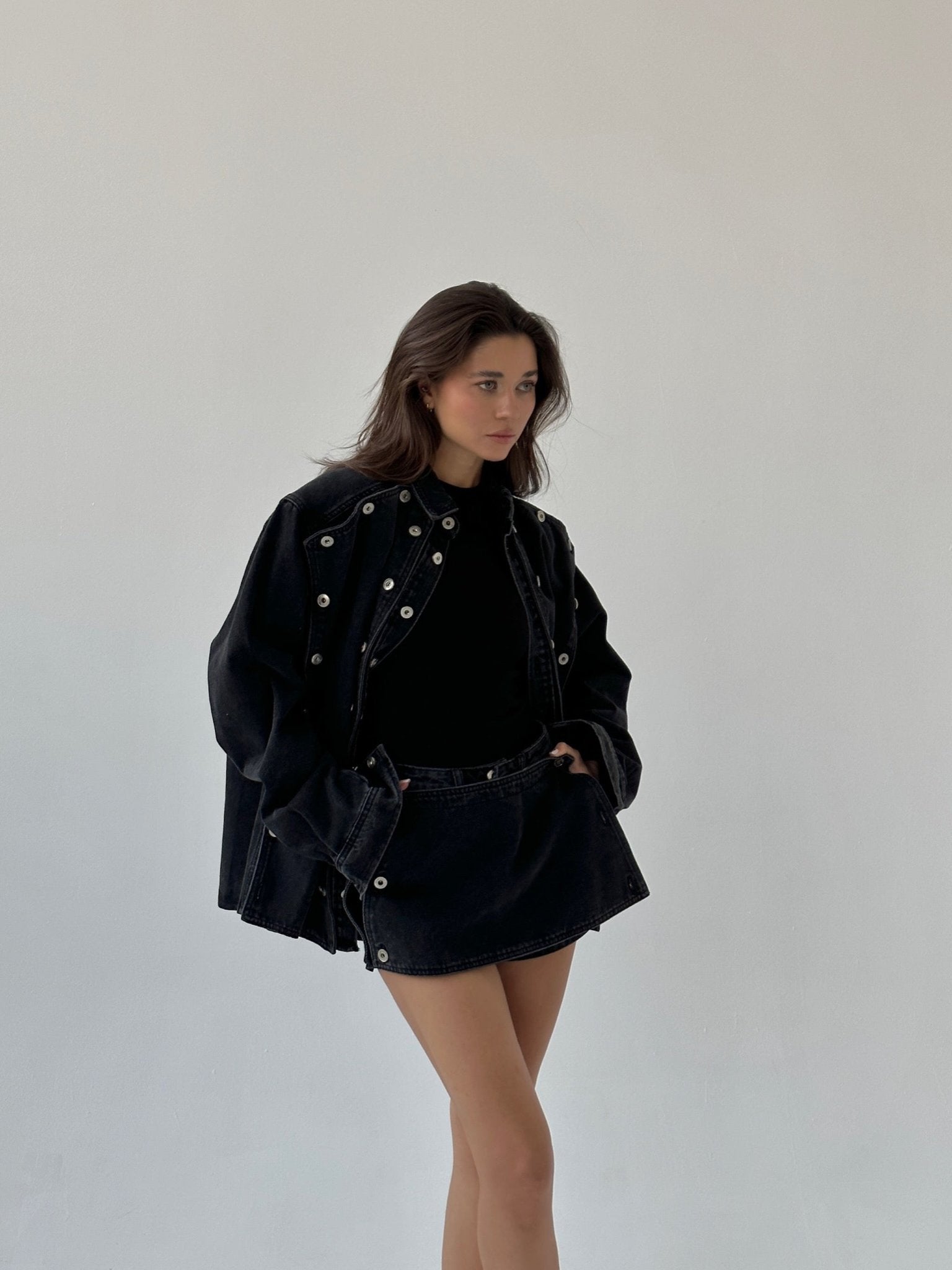 A person with long dark hair is posing against a plain background, wearing the Etta set. The ensemble features an oversized black button-up jacket adorned with silver grommets over a black top and matching short skirt. One hand is in their pocket, and they have a serious expression.