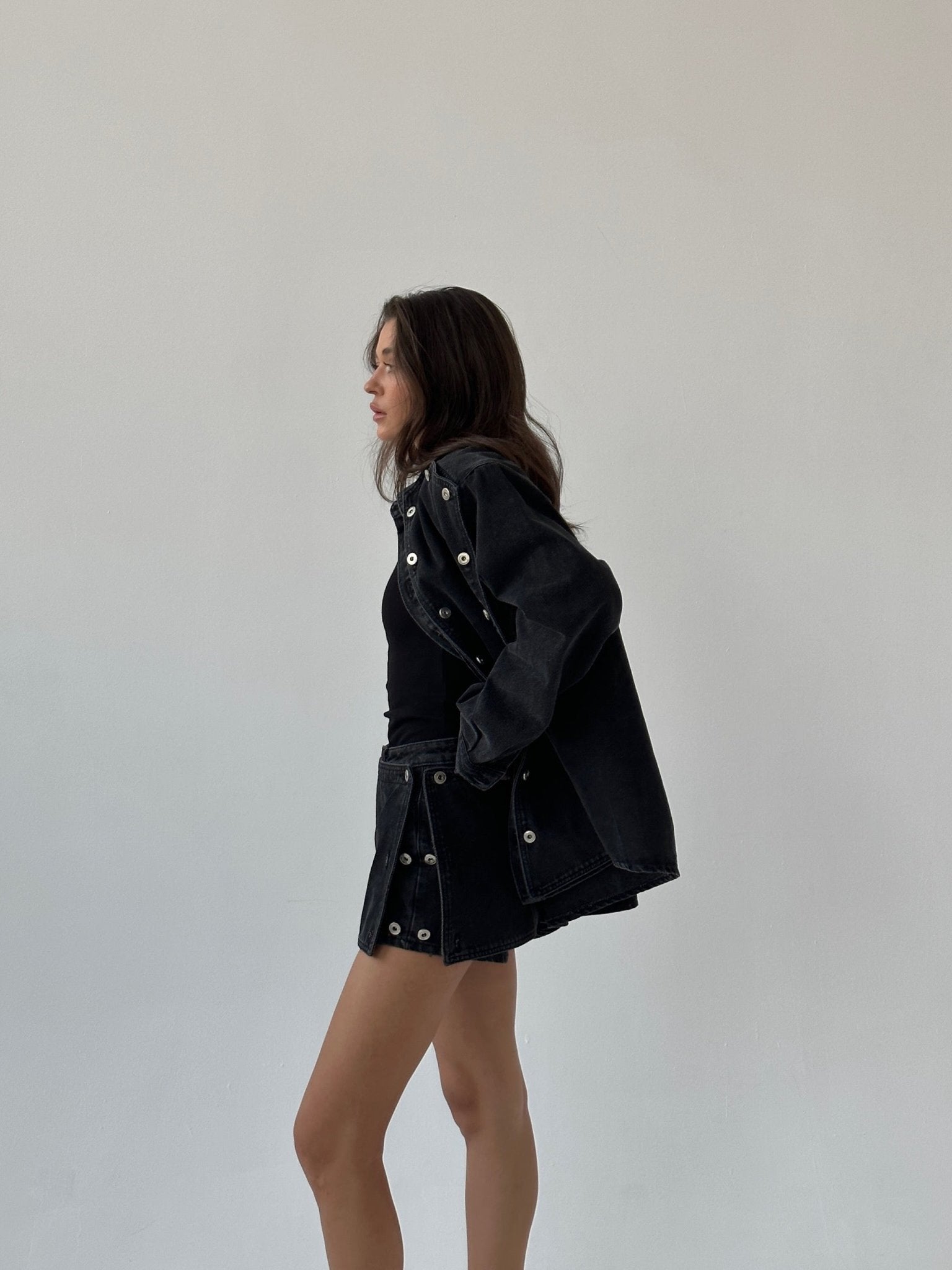 A woman stands sideways against a plain background, wearing the Etta set—a black denim outfit featuring a cropped jacket adorned with large silver grommets, paired with a matching skirt. She has her hand in her pocket and looks relaxed as her hair falls loosely over her shoulders.