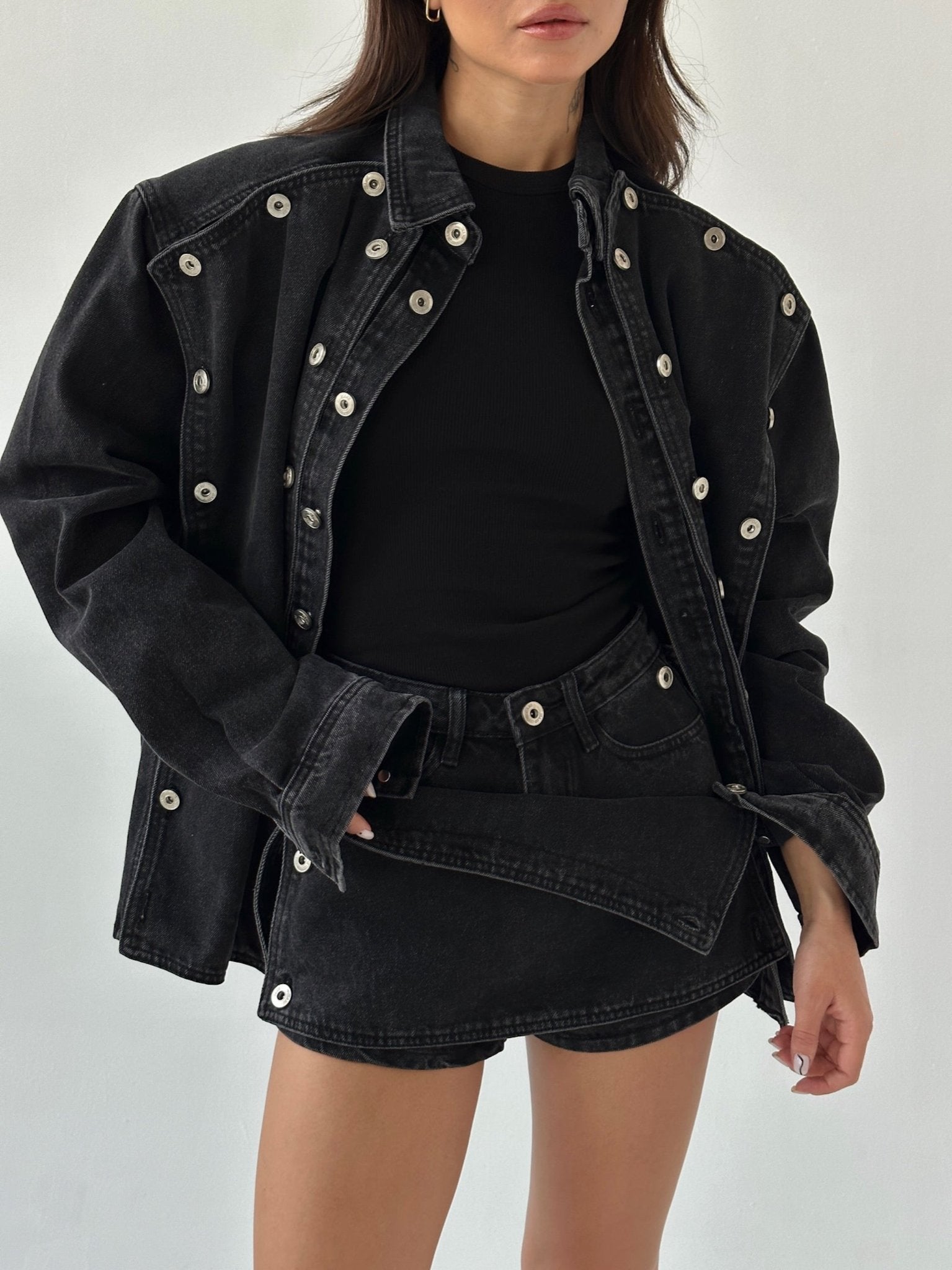 A person is wearing the Etta set, which includes an oversized black denim jacket adorned with silver grommets, a black top, and a matching denim mini skirt. The outfit exudes a casual and edgy vibe. The plain white background accentuates the fashion ensemble.