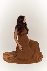 A woman with long, dark hair is seated on the floor in a side profile, wearing the Ezra dress— a sleeveless maxi dress with a waist-hugging silhouette that drapes elegantly around her. The background is a plain, light color. She has one hand resting on her lap and the other on her lower abdomen.