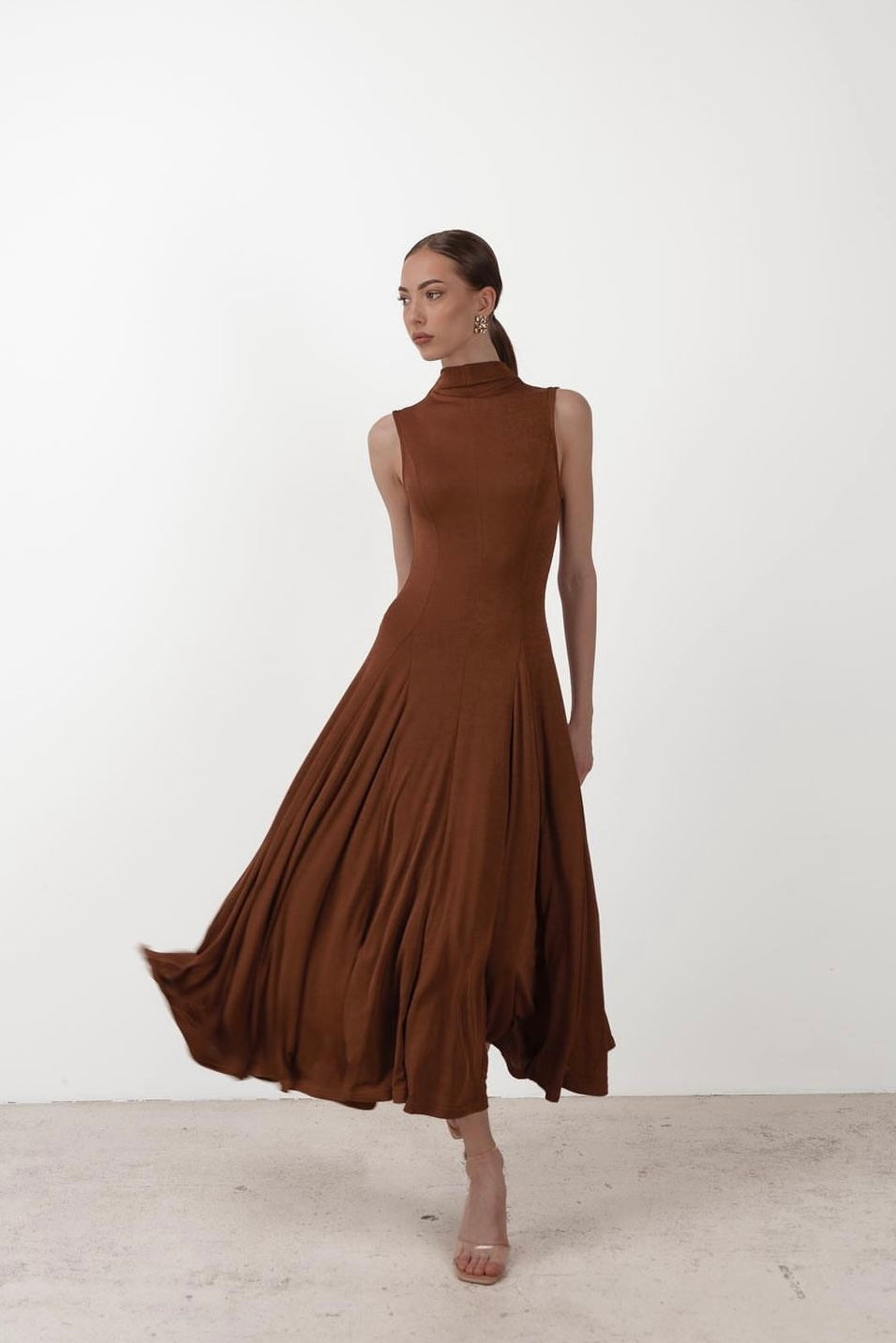 A woman in the Ezra dress, a sleeveless maxi, poses against a plain white background. With one leg slightly forward and her gaze directed to the side, she showcases the flowing brown fabric that elegantly flares out. The stretchable material of the dress and her clear high-heeled shoes complete her chic look.