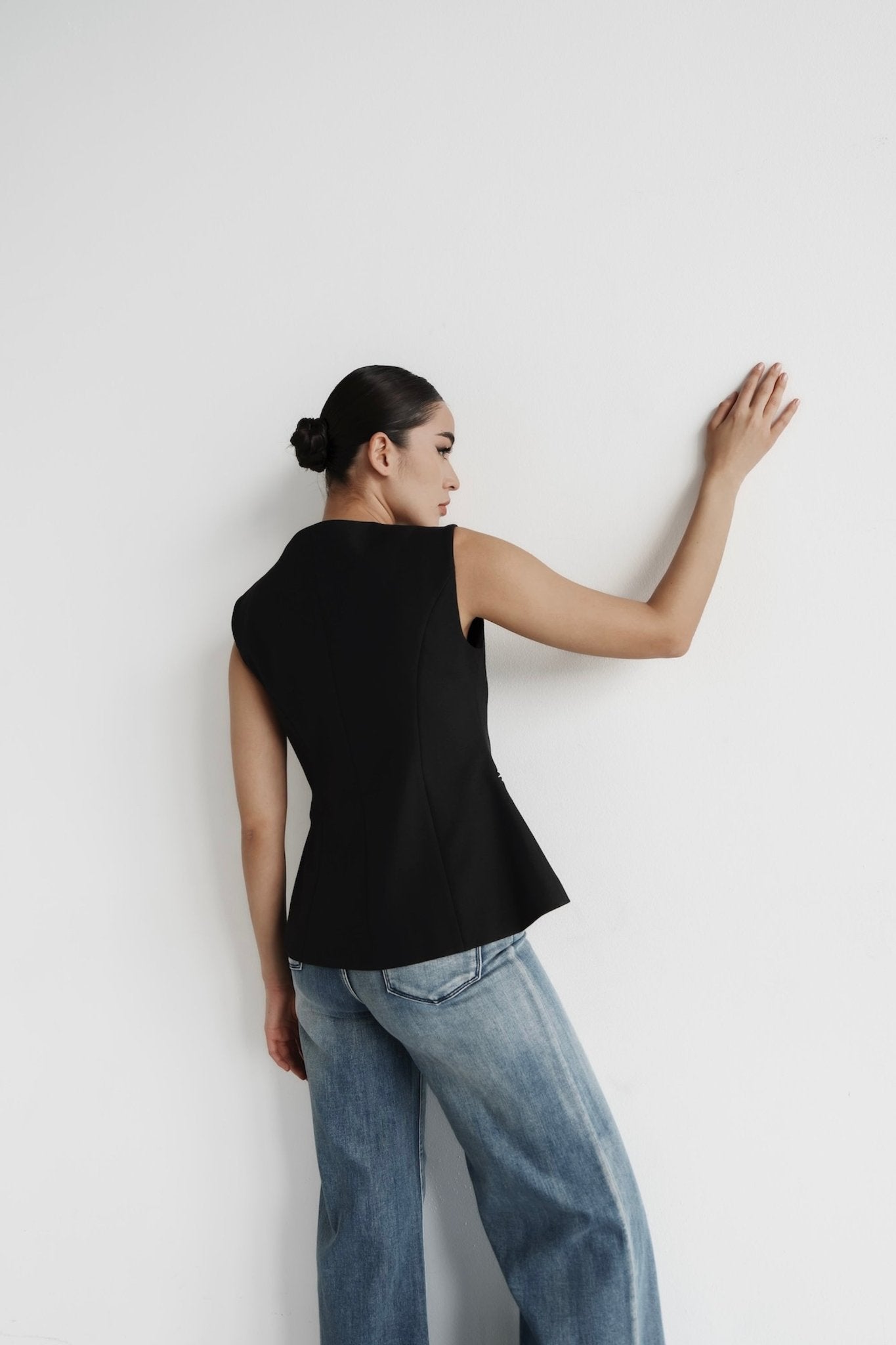 A person with long hair styled in a sleek bun stands against a white wall, facing away. They are wearing a sleeveless button-up black Eloise Vest, which features a flared hem, and light blue wide-leg jeans. Their left hand is placed on the wall while their right arm rests by their side, perfectly showcasing the timeless design of the Eloise Vest.