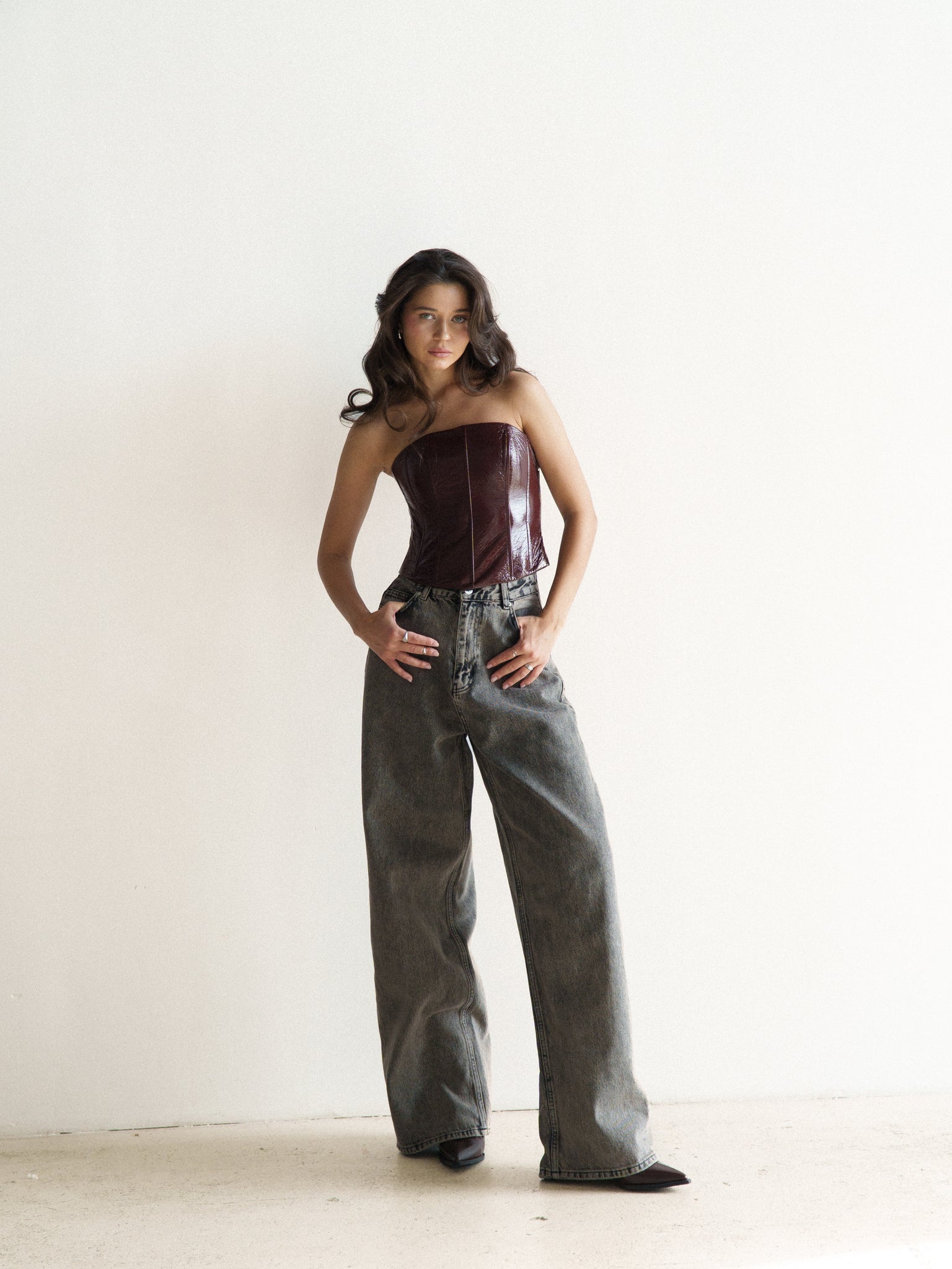 A woman stands against a plain white background, wearing an Imari Corset in deep burgundy paired with loose-fitting, high-waisted gray jeans. She has long, wavy brown hair cascading over her shoulders and is sporting black high-heeled boots. With her hands resting on her hips, this ensemble exudes timeless style and is an essential addition to any wardrobe.