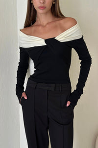 A person stands against a light-colored wall, wearing the Amélie knit top with off-the-shoulder design and white fabric around the neckline. Their hands are in the pockets of their high-waisted black pants. The outfit is a versatile piece that exudes a sophisticated look, effortlessly stylish and elegant.