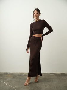 In a minimalist setting, a woman exudes modern femininity as she confidently poses in the Gaïa Set. The ensemble features a dark brown ribbed long-sleeve crop top and matching skirt, both expertly crafted from high-quality cotton knit fabric. Her look is effortlessly completed with an elegant loose updo and beige heels, set against a plain white wall.