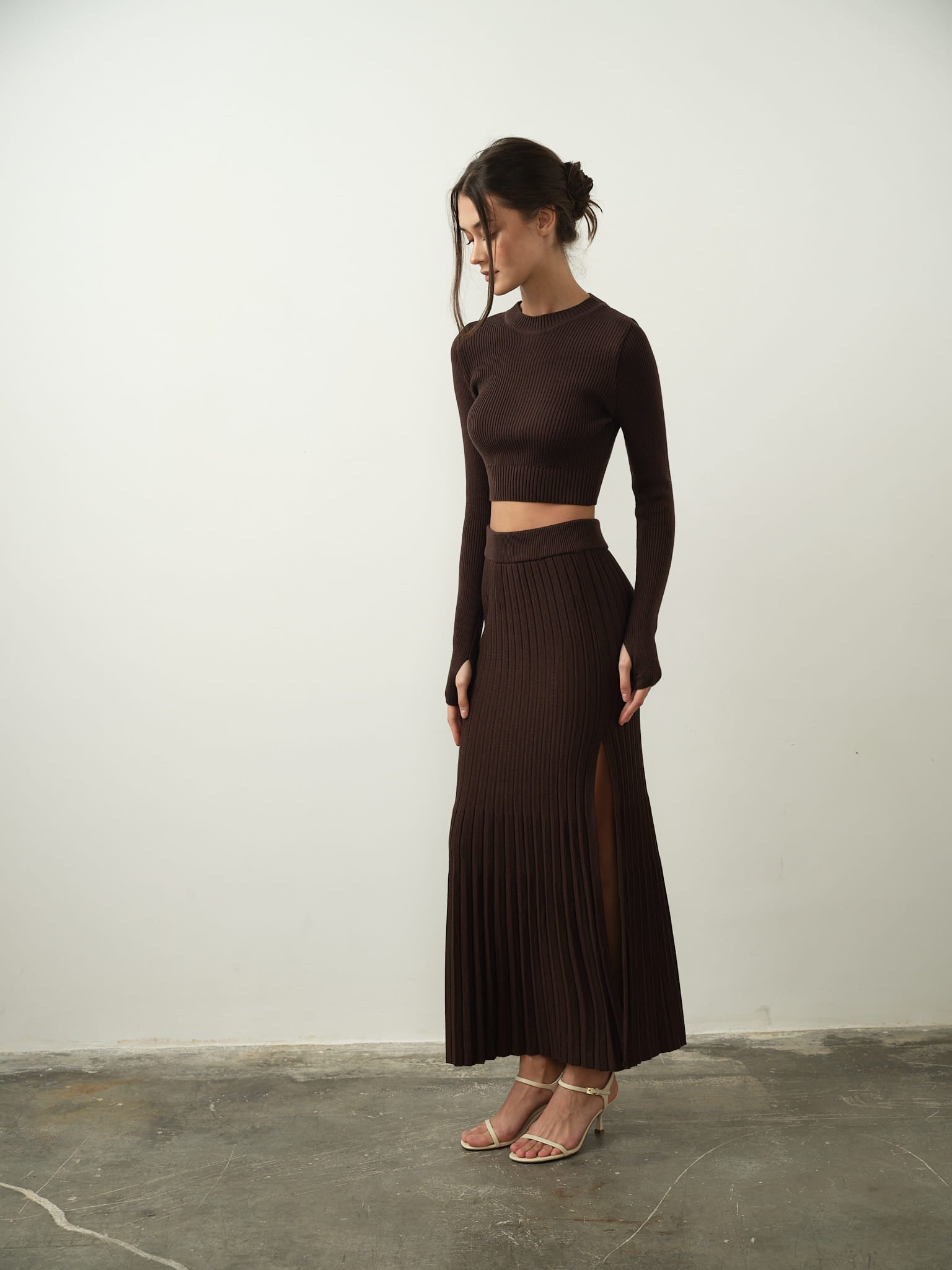 A woman dressed in the Gaïa Set stands on a plain floor against a neutral wall, exuding modern femininity. The set includes a cropped brown long-sleeve top paired with a pleated maxi skirt. She has her hair tied back and her arms relaxed, complementing the outfit with strappy beige heels. The ensemble highlights its high-quality cotton knit fabric.