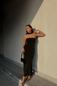 A woman in a black Hailey dress, which is figure-hugging and strapless, paired with heels, stands against a wall where shadow play is evident. She holds a black shoulder bag and gazes slightly off-camera as the setting sun casts intriguing light and shadow patterns on the wall behind her.