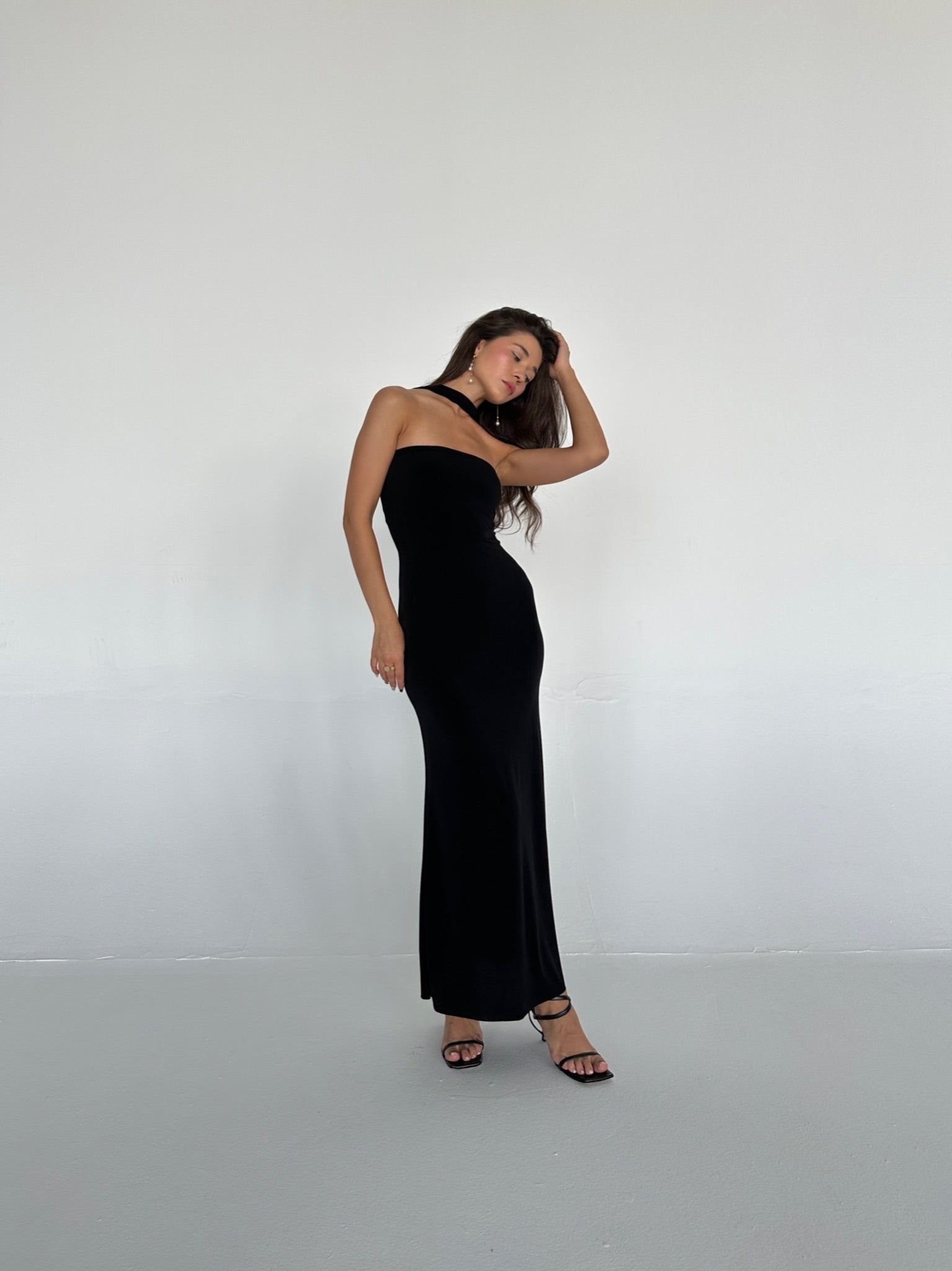 A woman is standing in a minimalist setting, wearing the floor-length, figure-hugging Hailey dress with a slit. She has one hand resting on her forehead and the other on her hip, with long hair cascading over one shoulder, and is wearing black high heels.