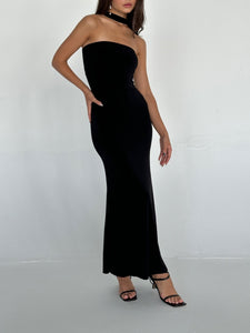 A person wearing a figure-hugging, strapless Hailey dress in black poses against a plain white wall. They are also wearing black high-heeled sandals. One hand is on their hip, and the other arm is relaxed by their side.