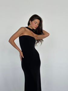 A woman with long, wavy hair is posing against a plain white background. She is wearing the Hailey dress, a black figure-hugging strapless outfit featuring a high neck choker detail. Her left hand is placed on her hip while her right hand touches her hair. She has a serene expression.