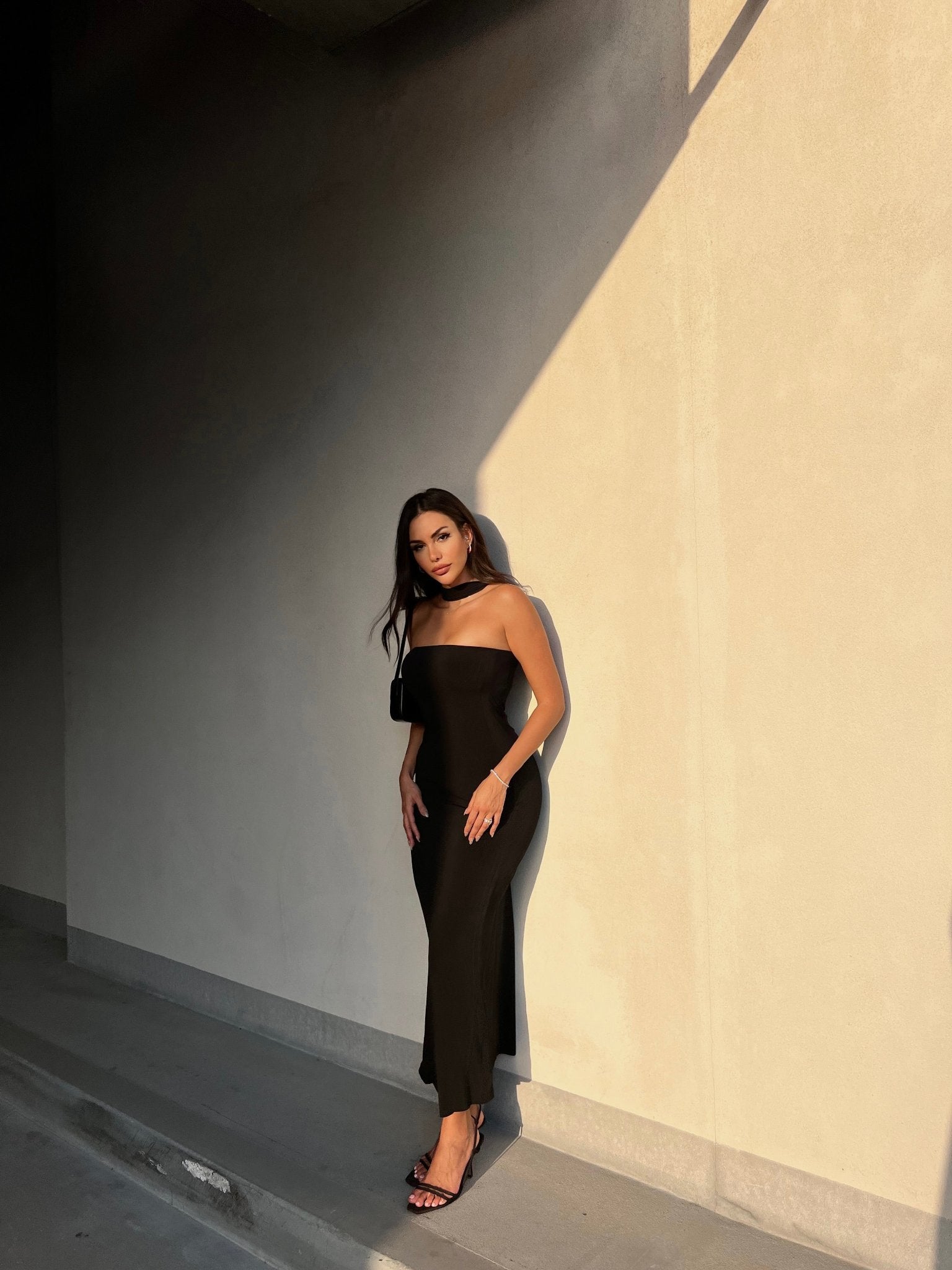 A woman with long dark hair wearing the strapless, figure-hugging Hailey dress in black and matching black heels is standing against a light-textured wall. Sunlight casts a dramatic shadow behind her, creating a striking contrast. She has one hand resting on the wall and is looking at the camera.