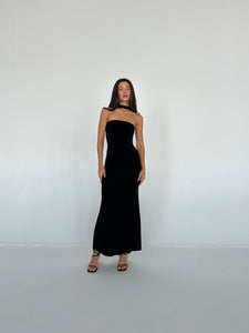 A woman stands against a plain light-colored wall, wearing the Hailey dress that fits closely to her figure. She has long, dark hair and is wearing black high-heeled sandals. Her arms are relaxed by her sides, and she is looking directly at the camera.