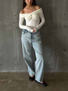 A person is standing against a concrete wall wearing the Sabella top—an elegant white off-the-shoulder piece with long sleeves and a twist detail, crafted from high-quality soft cotton fabric. The look is paired with light-wash high-waisted jeans and black ballet flats. The person has long hair and is holding their hands together loosely.