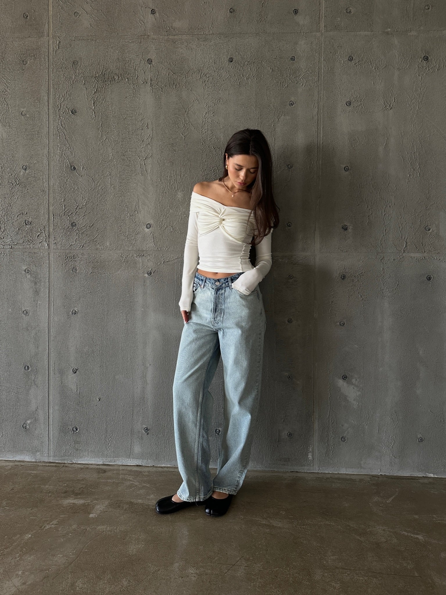 A woman with long dark hair stands against a textured gray wall. She wears the Sabella top, an off-the-shoulder white piece made of high-quality soft cotton fabric, paired with light blue wide-legged jeans and black shoes. The woman gazes downward, with her hands loosely placed in her pockets.