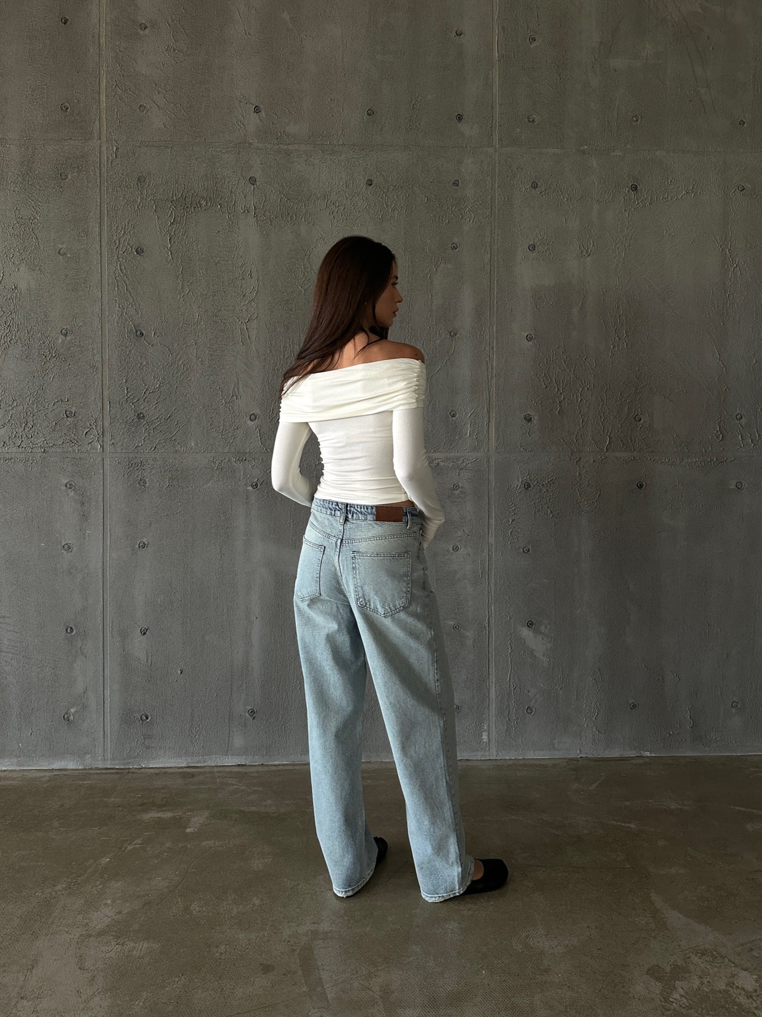 A person with long hair stands facing a textured, gray wall. They are wearing the Sabella top, an off-the-shoulder white blouse made from high-quality soft cotton fabric, and loose-fitting light blue jeans with hands in their pockets. The person is also wearing black shoes, and the floor appears to be concrete.