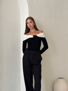 A woman with long hair is standing against a textured, light-colored wall in a minimalist setting. She is wearing the Amélie knit top and black high-waisted pants, creating a sophisticated look. A small, white ceramic vase is placed on the floor to her right.