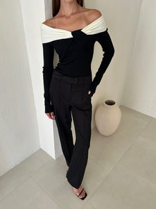 A woman is standing indoors wearing a stylish Amélie knit top, black high-waisted trousers, and black sandals. Posed near a white wall with a beige vase on the floor beside her, she exudes a sophisticated look against the minimal and modern background.