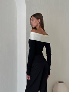 A woman with long brown hair stands with her back to the camera, facing slightly to the right. She is wearing an Amélie knit top, a black off-the-shoulder design featuring a white band across the shoulders, paired with black pants that exude a sophisticated look. Positioned next to a textured white wall, she stands by a simple round vase.