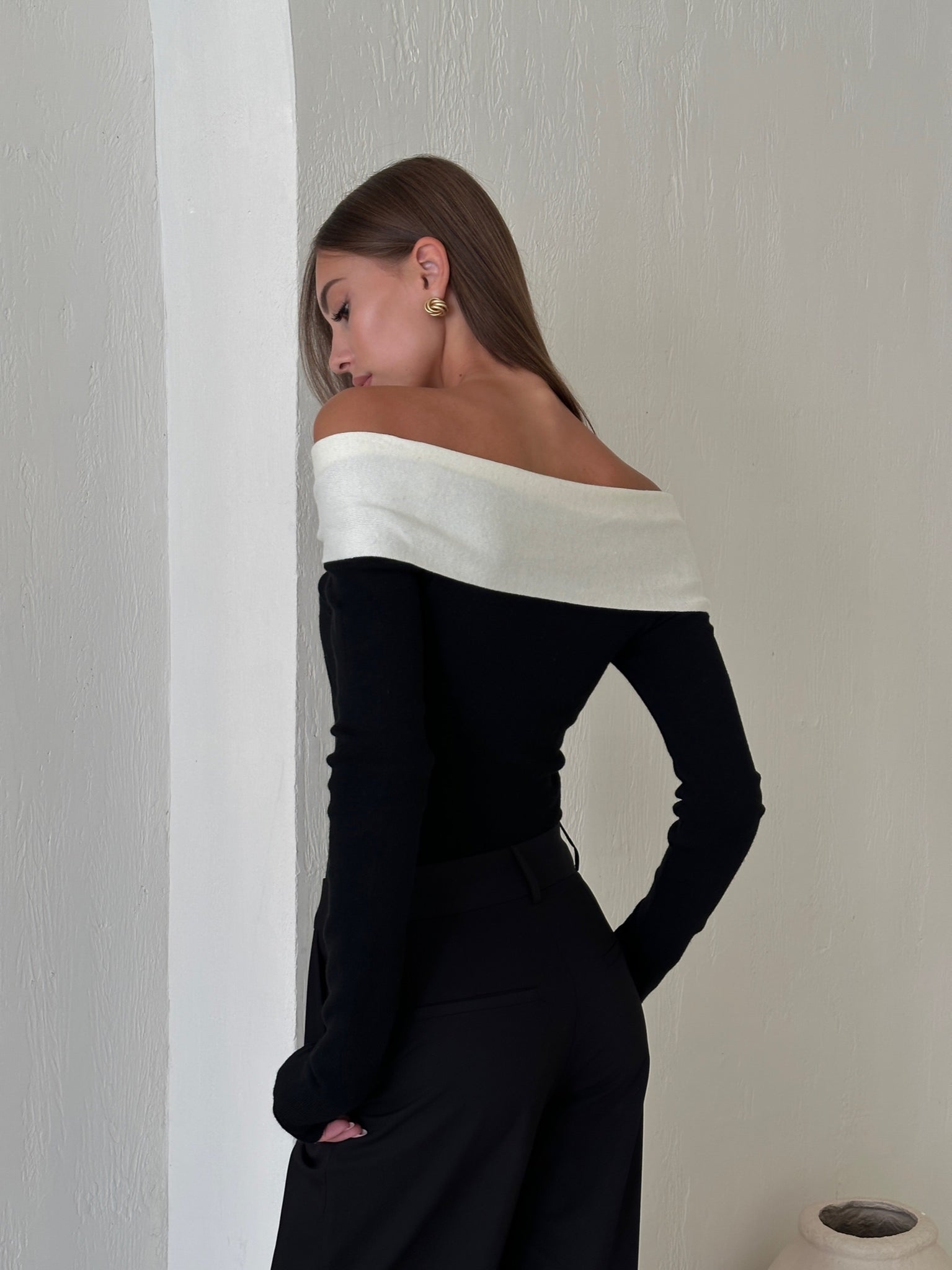 A woman with long brown hair stands sideways in a minimalist setting, wearing the Amélie knit top with an off-the-shoulder design and white neckline, paired with black pants. She accessorizes with gold earrings while facing a textured white wall. A ceramic vase is visible in the background, completing her sophisticated look.