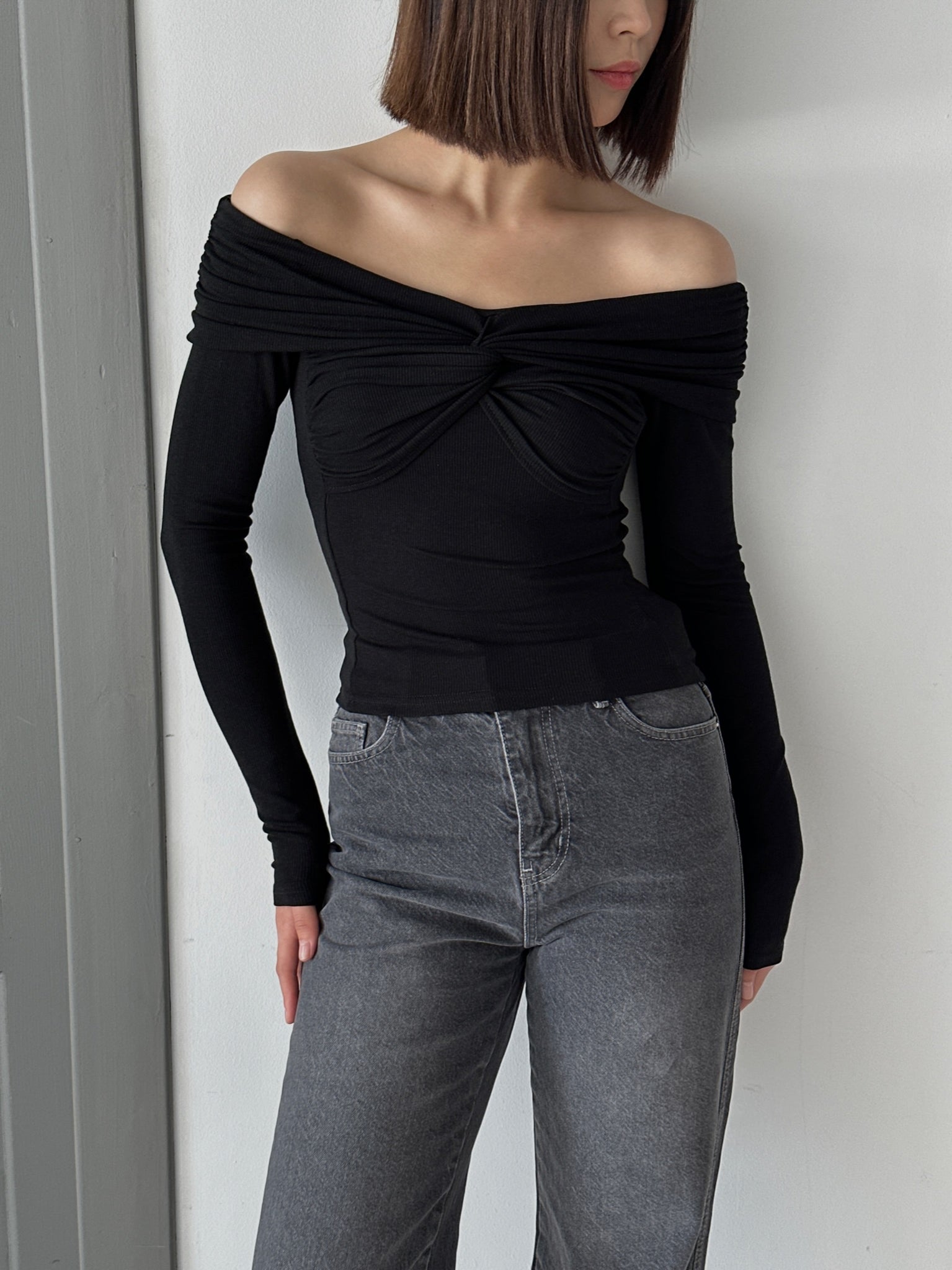 A woman with shoulder-length hair is wearing the Sabella top, an off-the-shoulder black garment made from high-quality soft cotton fabric, paired with high-waisted gray jeans. She stands in front of a plain wall, gazing slightly downward and to the side.