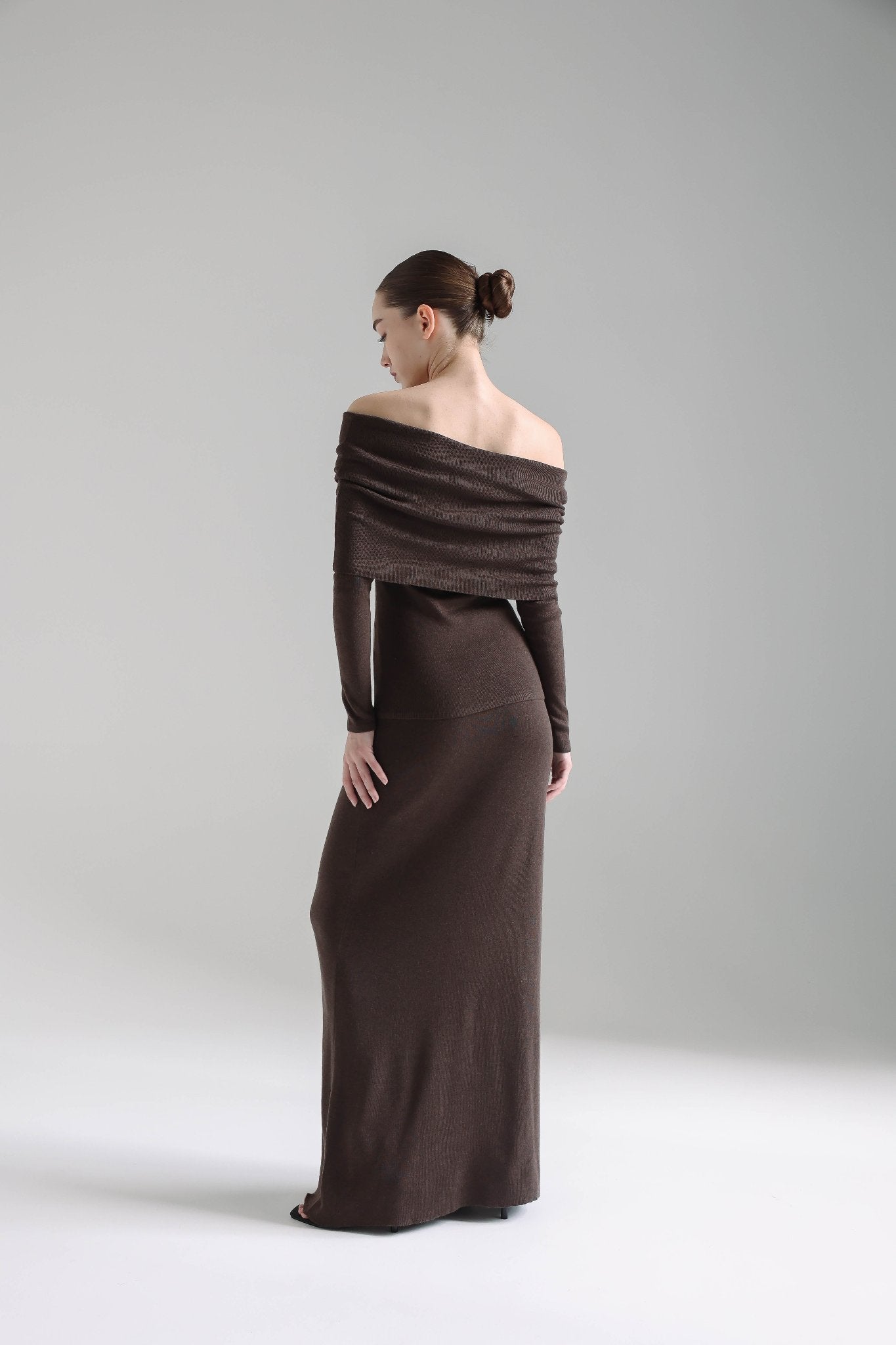 A person stands gracefully while facing away from the camera, elegantly draped in the Madeline knit set. The dark brown, off-the-shoulder ensemble with long sleeves exudes timeless sophistication. Their dark hair is neatly styled in a low bun, perfectly complementing the minimalist and elegant gray background.