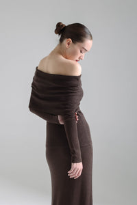 A person with their hair in a bun stands against a minimalist grey background. They are wearing the Madeline knit set in brown and are gazing downward with their left arm gently resting on their waist. The long sleeves of this versatile off-shoulder knitwear extend past the wrists, exuding timeless elegance.