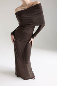 A person is standing against a plain background, wearing the Madeline knit set, which features a long, off-the-shoulder design in dark brown with long sleeves, exuding timeless elegance. One hand rests on their hip while the other arm hangs by their side. Their face is not visible in the image.