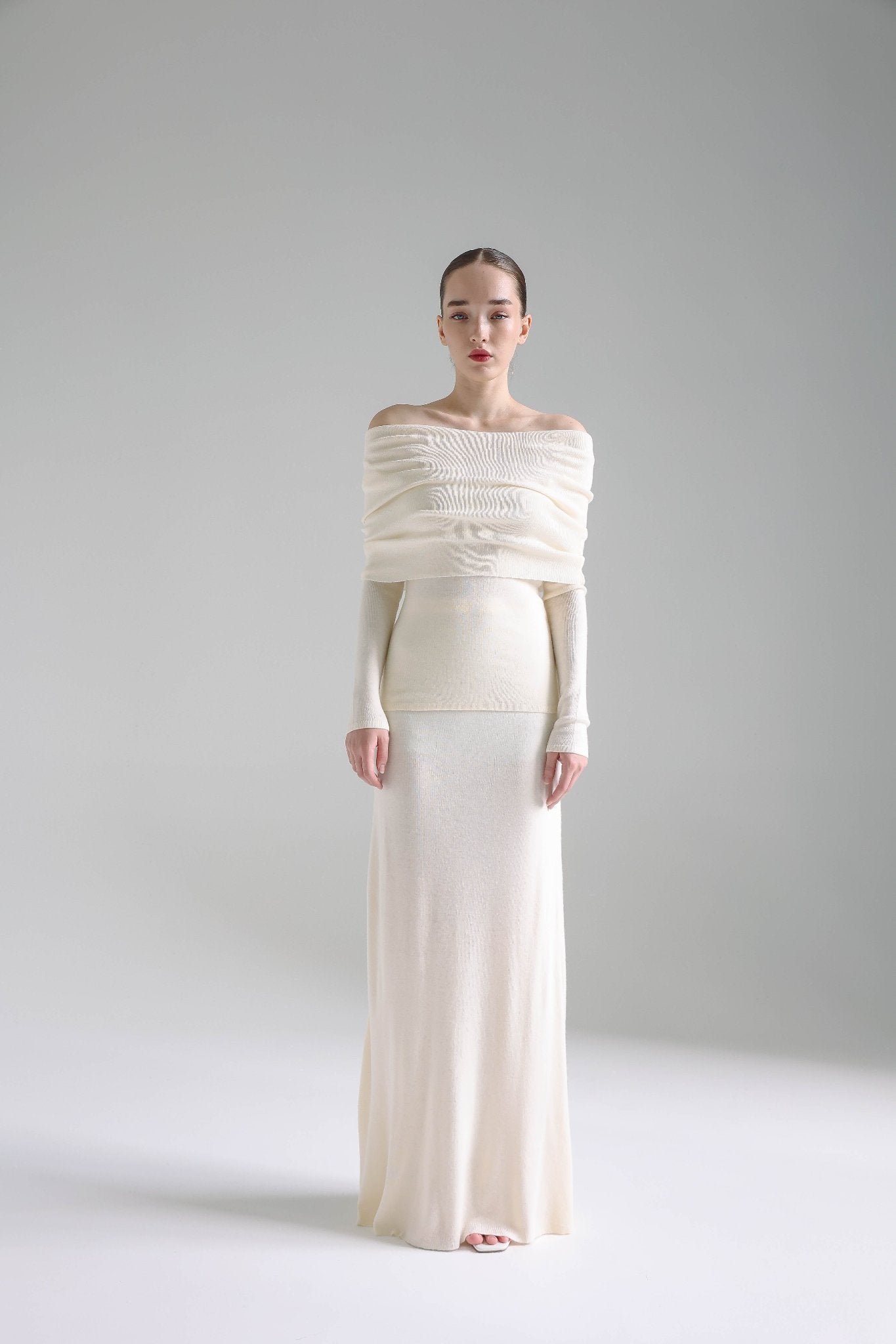 A woman stands against a plain background wearing the Madeline knit set, featuring a long, off-shoulder, cream-colored dress with long sleeves. Her hair is slicked back, and she faces forward with a neutral expression. The overall setting exudes timeless elegance and is minimalist in its approach.