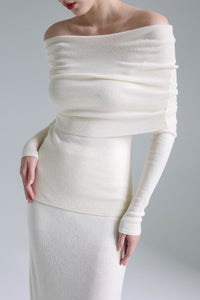 A person wearing a sophisticated Madeline knit set, characterized by an off-the-shoulder design and long sleeves in creamy white, stands against a plain, light gray background. The outfit showcases a draped neckline and a fitted silhouette that exudes timeless elegance while highlighting the person's figure.