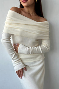 A person is wearing the Madeline knit set, featuring an off-the-shoulder cream-colored ribbed knit sweater with a matching skirt. Showcasing timeless elegance, the long-sleeve knitwear elegantly drapes across the shoulders, covering the hands. The individual stands against a plain, light-colored background.