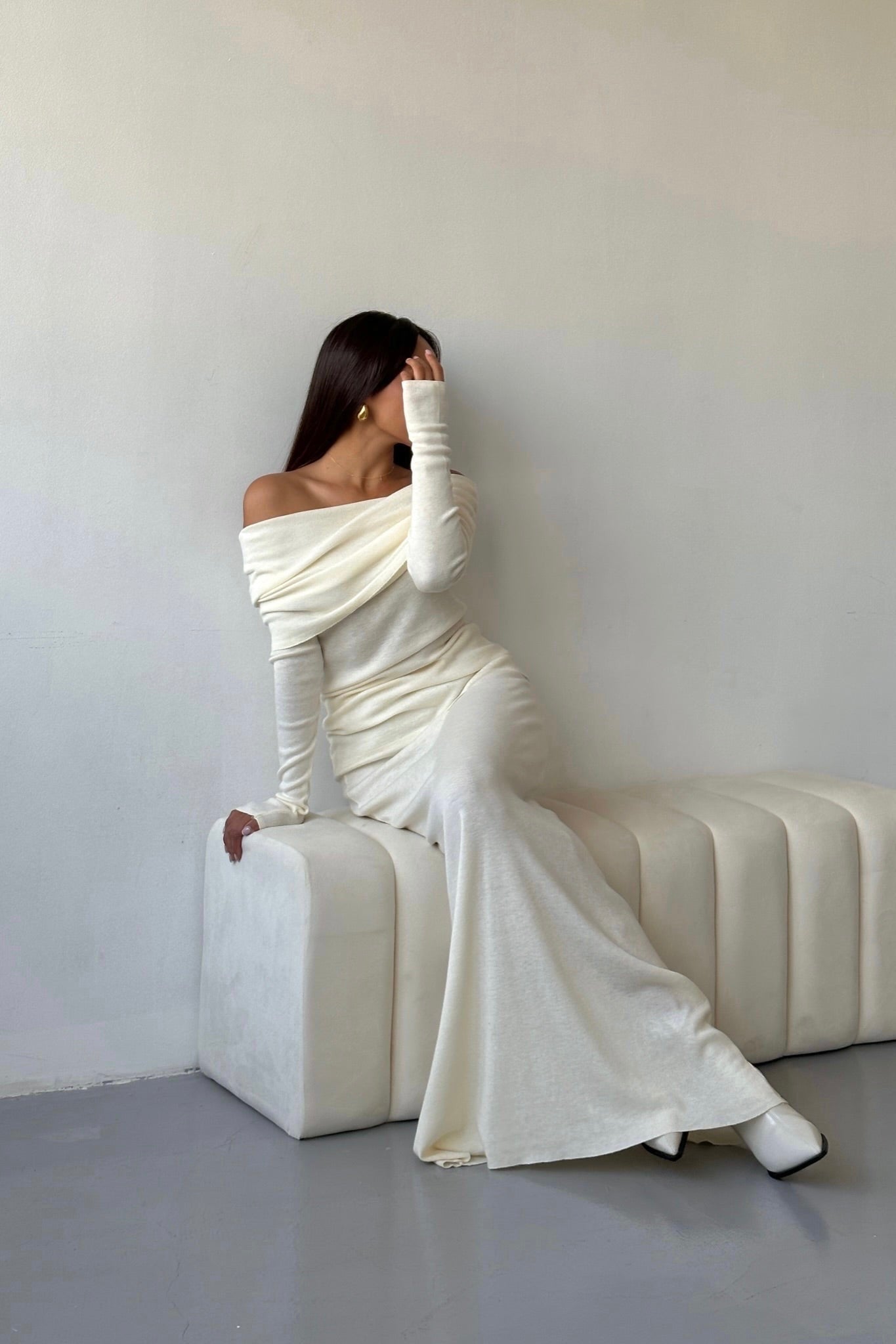 A woman with timeless elegance covers part of her face with her hand as she sits on a white cushioned bench. She is wearing the Madeline knit set, an off-the-shoulder, long-sleeved, floor-length white gown complemented by matching shoes. The background features a plain light-colored wall and floor.