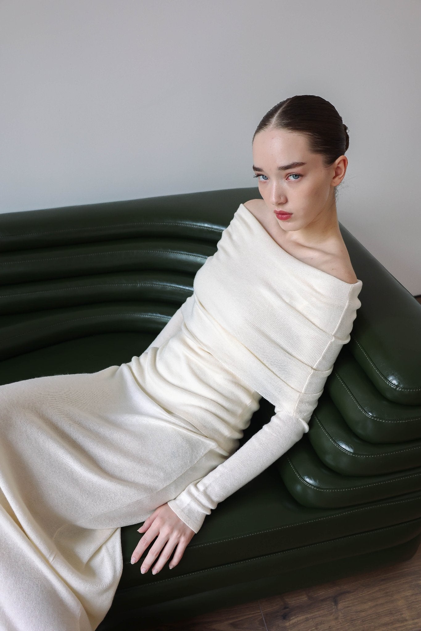 A woman with dark hair is sitting on a modern, green leather couch. She is wearing the Madeline knit set, an off-shoulder, long-sleeved white dress from the latest collection. The background is plain and minimalistic, and she exudes timeless elegance with her calm, composed expression.